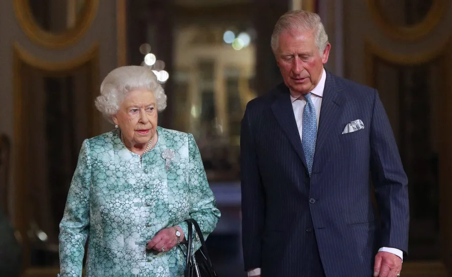 Two older individuals in formal attire walking together indoors, engaged in conversation.