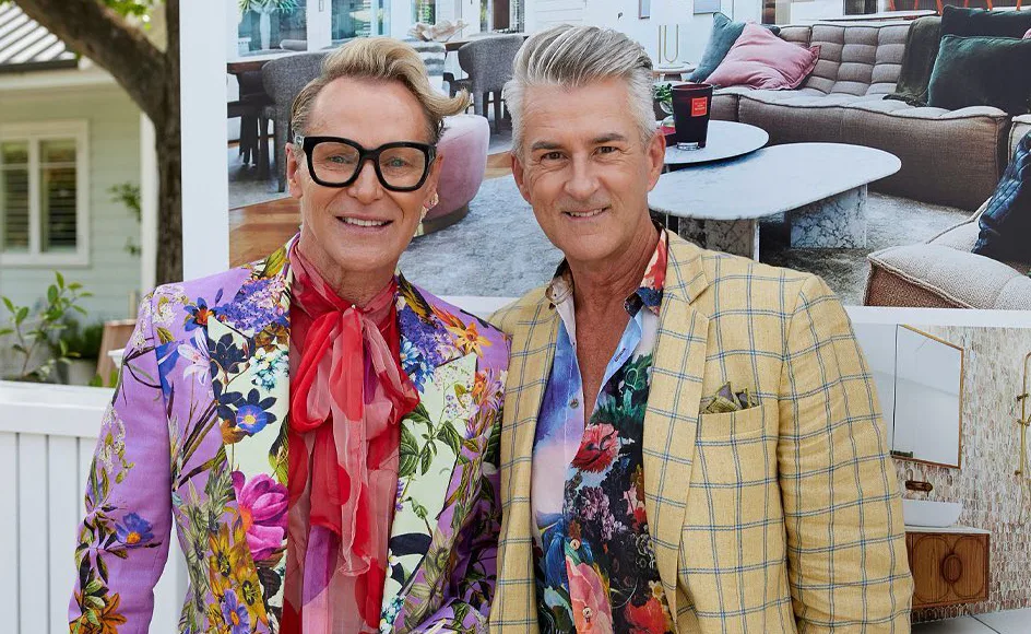 Two men smiling, wearing floral jackets and glasses, standing outdoors with a modern interior photo in the background.