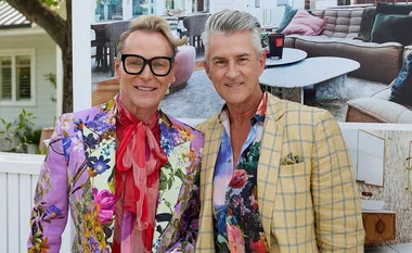 Two men smiling, wearing floral jackets and glasses, standing outdoors with a modern interior photo in the background.