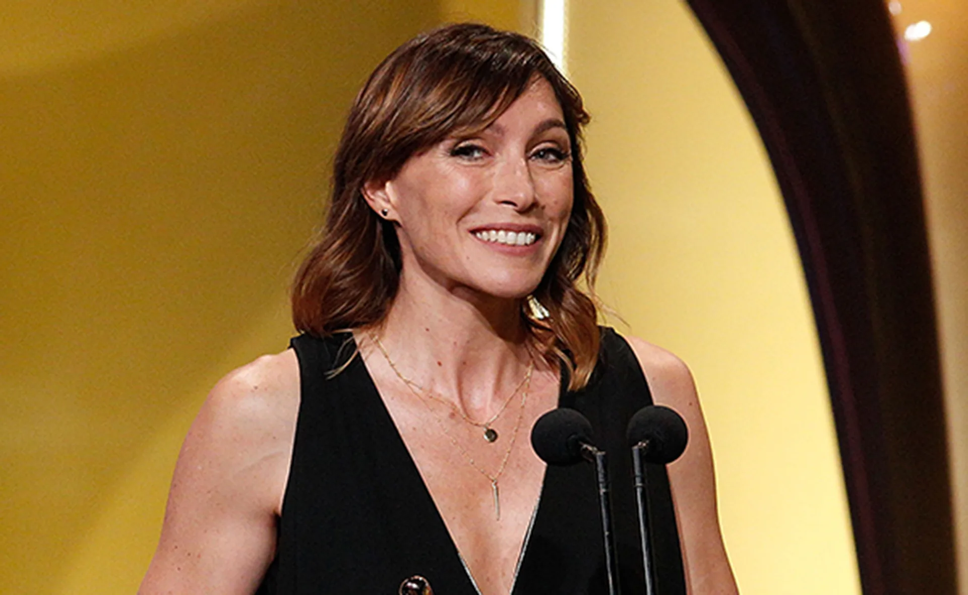 A woman in a black dress speaks into a microphone on stage with a gold background.