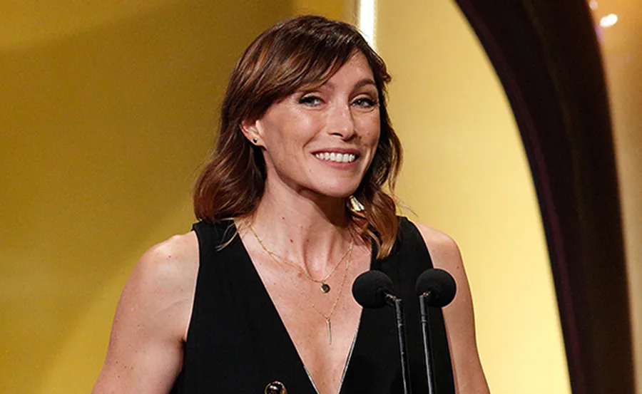 A woman in a black dress speaks into a microphone on stage with a gold background.