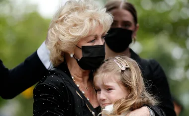 Elderly woman in a mask hugs a young girl at a funeral, expressing emotional support.