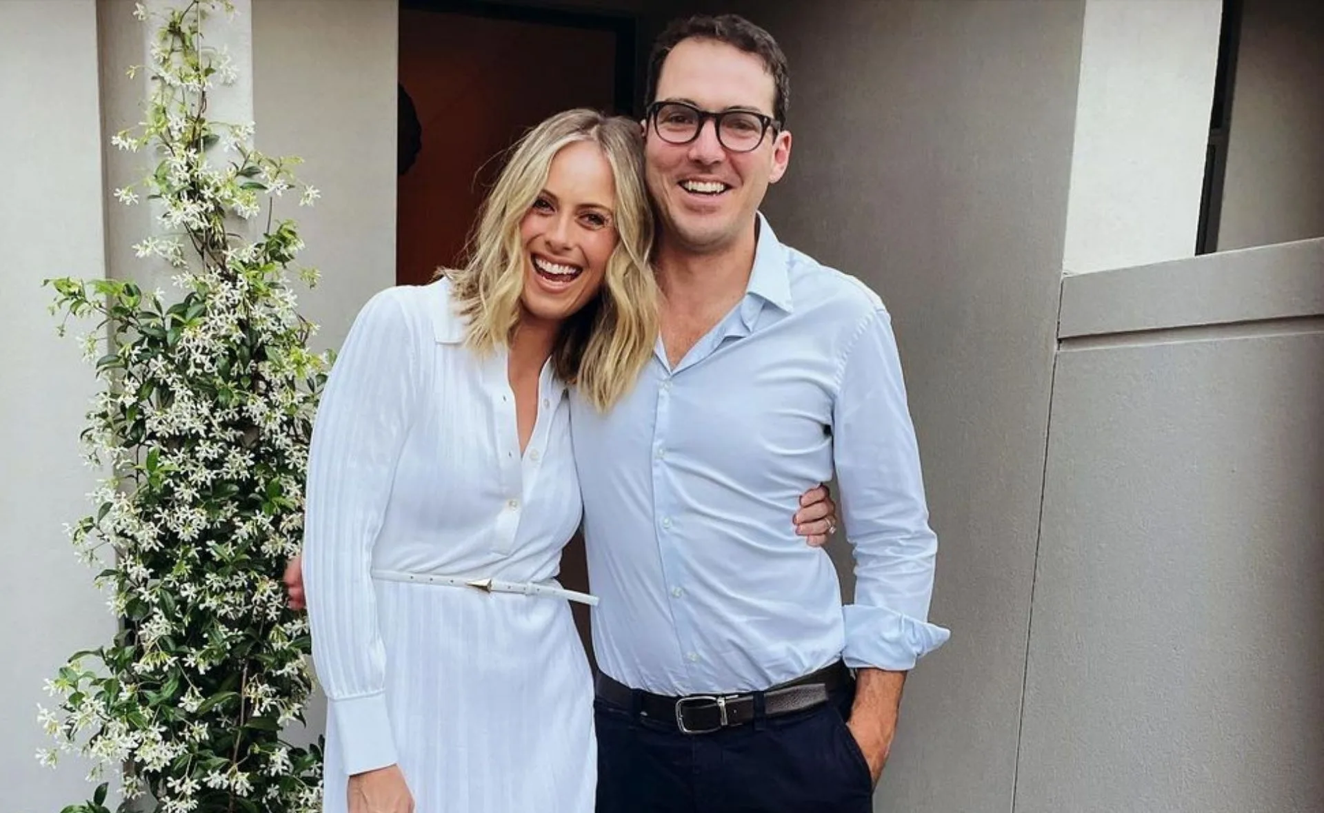 Couple smiling, posing together outdoors with a flowering plant on the side. Both are dressed in light-colored outfits.