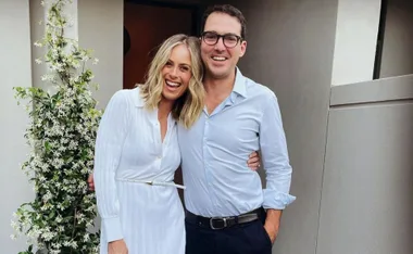 Couple smiling, posing together outdoors with a flowering plant on the side. Both are dressed in light-colored outfits.