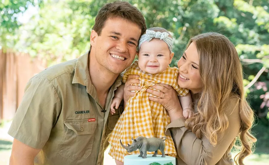 Family of three smiling outdoors, baby in a yellow dress, with a cake featuring a rhino figurine in the foreground.