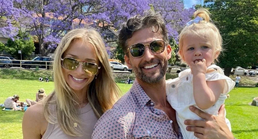 Family outdoors at park, woman in sunglasses, man holding blonde toddler.
