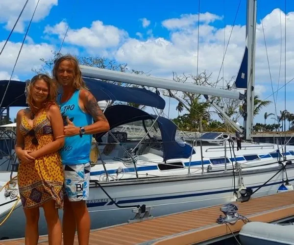 Couple standing on a dock in front of a white sailboat under a blue sky.