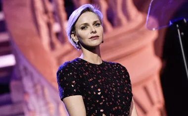 Blonde woman in a black dress with red accents, standing in front of an ornate background.
