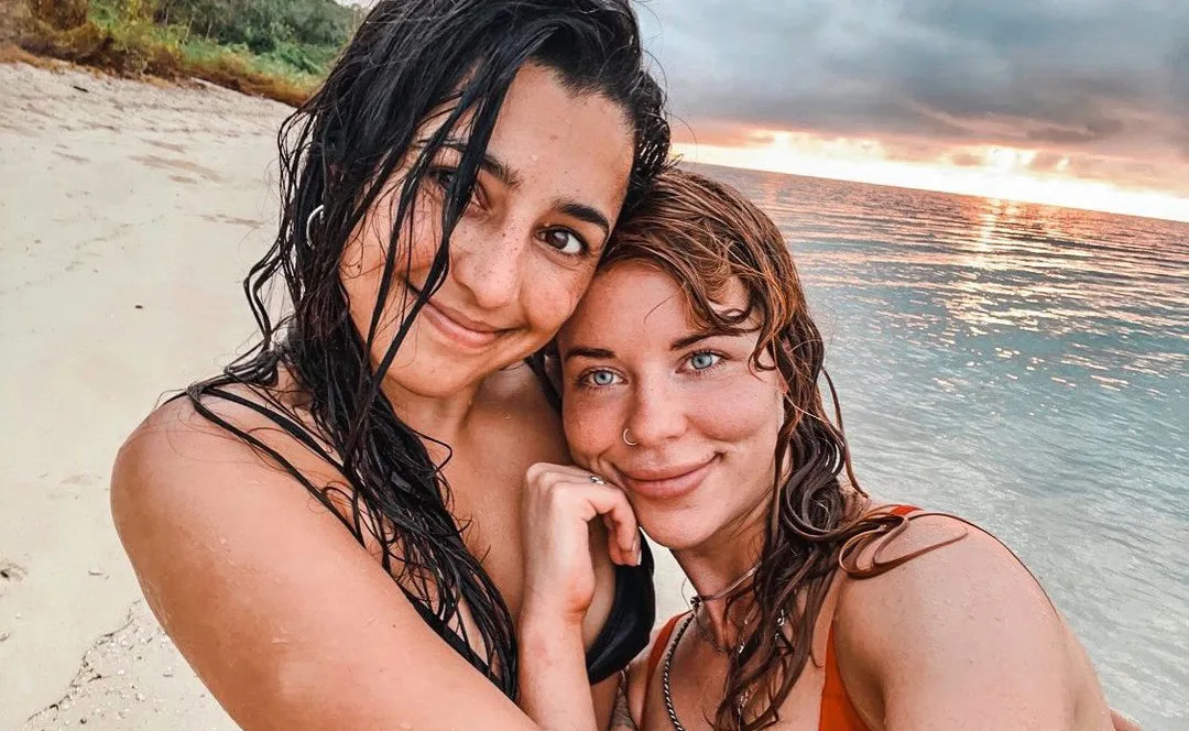 Two women with wet hair smile on a beach at sunset, ocean waves in the background.
