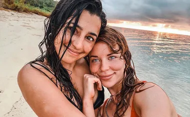 Two women with wet hair smile on a beach at sunset, ocean waves in the background.