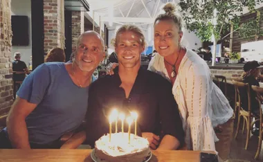 Three people smiling at a table with a lit birthday cake in a restaurant setting.