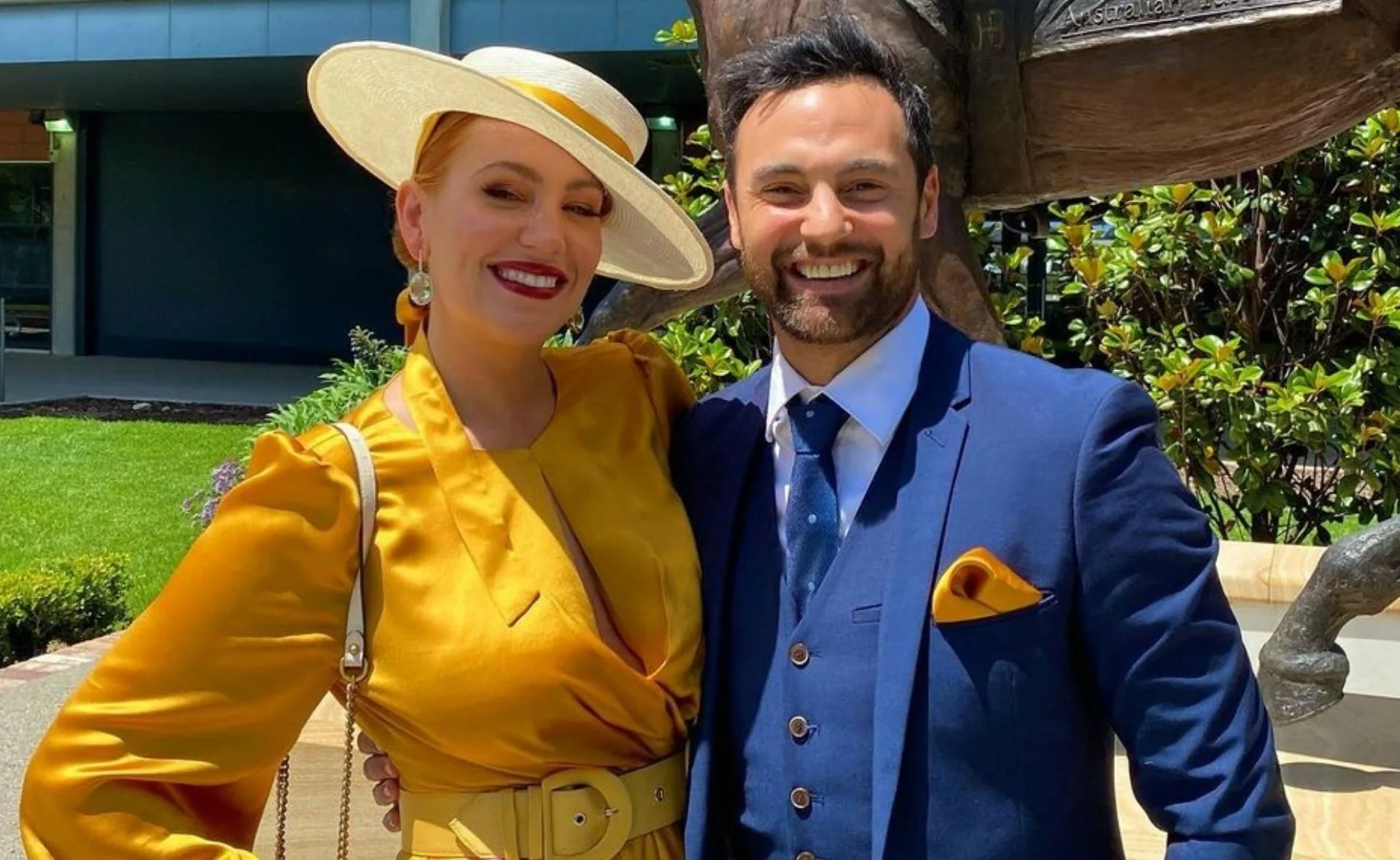 A couple smiling, dressed formally; the woman in a yellow dress and hat, and the man in a blue suit, outdoors.