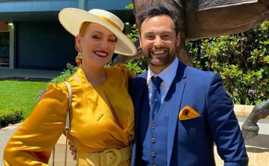 A couple smiling, dressed formally; the woman in a yellow dress and hat, and the man in a blue suit, outdoors.