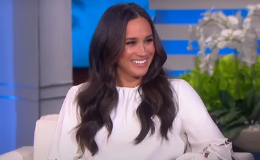 A woman with long dark hair in a white top smiles while seated on a talk show set with plants in the background.