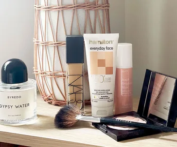 A collection of skincare and makeup products including sunscreen, foundation, and a makeup brush on a wooden table.
