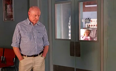 Man in checkered shirt looks somber outside hospital room where a woman lies in bed.