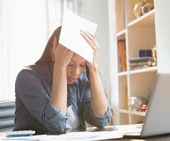 Woman stressed over finances, holding bills at a desk with a laptop.