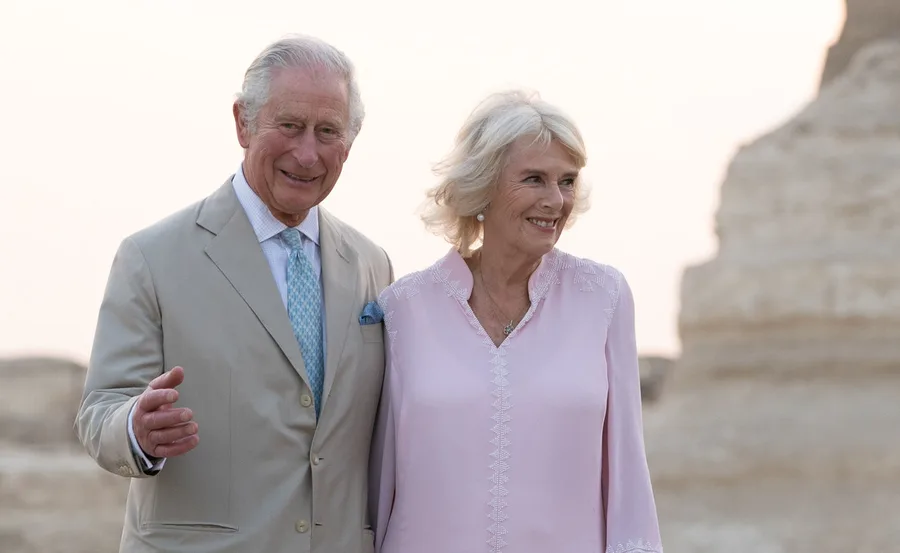 Royal couple smiling outdoors, dressed formally, against a rocky background.