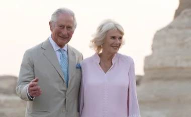 Royal couple smiling outdoors, dressed formally, against a rocky background.