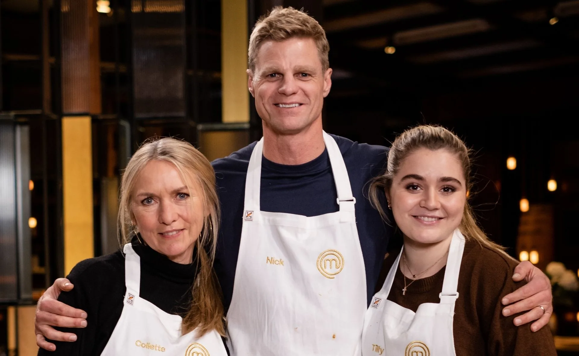 Three people wearing aprons with the MasterChef logo, smiling and posing together in a dimly lit environment.