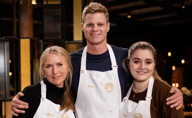 Three people wearing aprons with the MasterChef logo, smiling and posing together in a dimly lit environment.