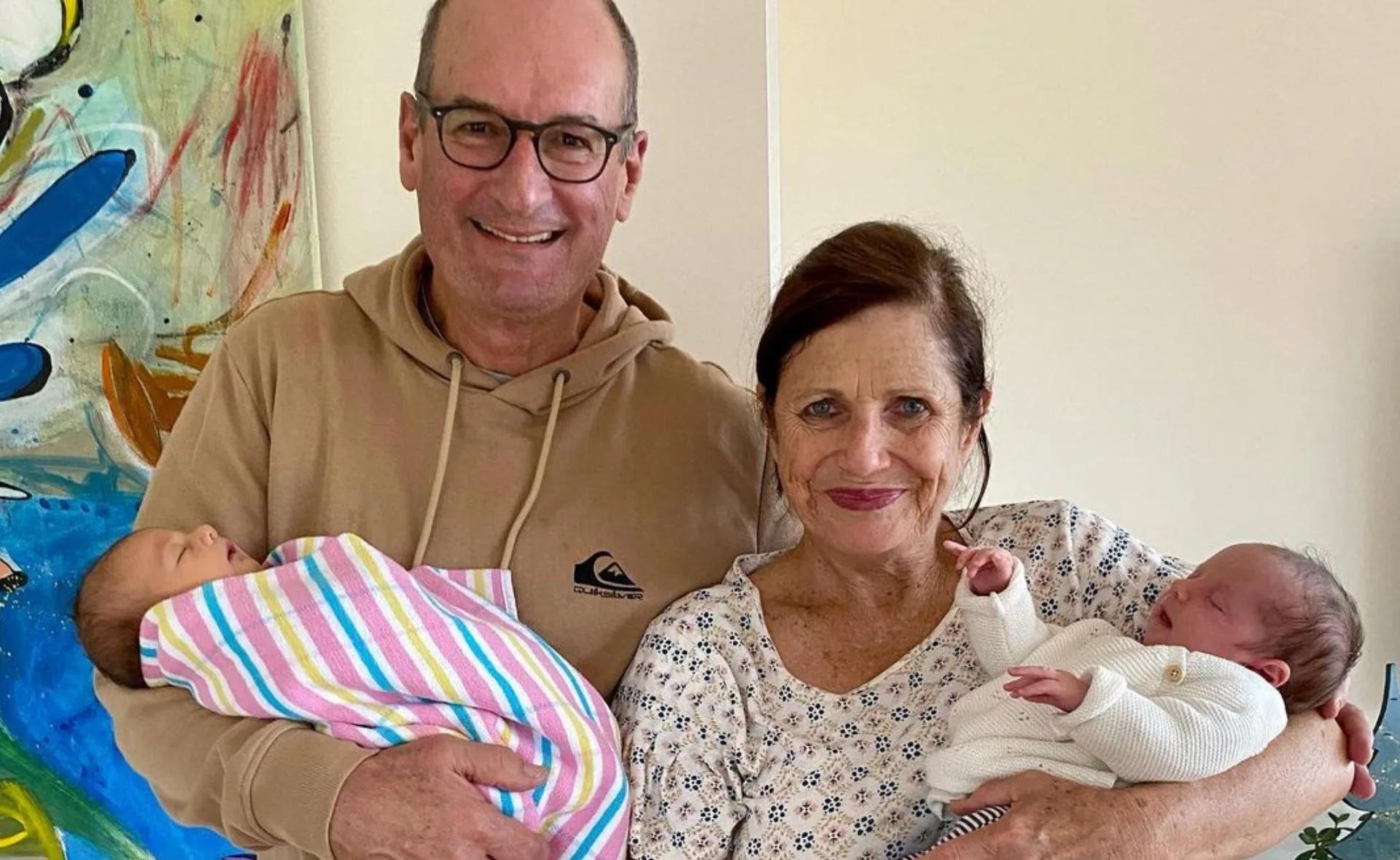 Grandparents smiling while holding two newborns wrapped in blankets, standing in front of colorful artwork.