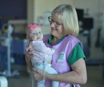 Volunteer woman in pink vest holding a baby with a headband and tube in a hospital setting.