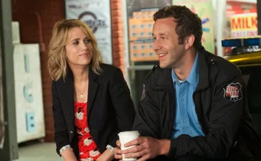 A woman in a blazer and a man in a uniform sit outside, smiling and talking while holding coffee cups.