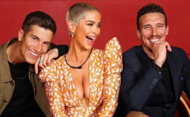 Three people smiling and posing against a red background, one wearing an ornate orange dress.