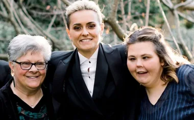 Three people smiling outdoors, one in a suit with two others on either side.