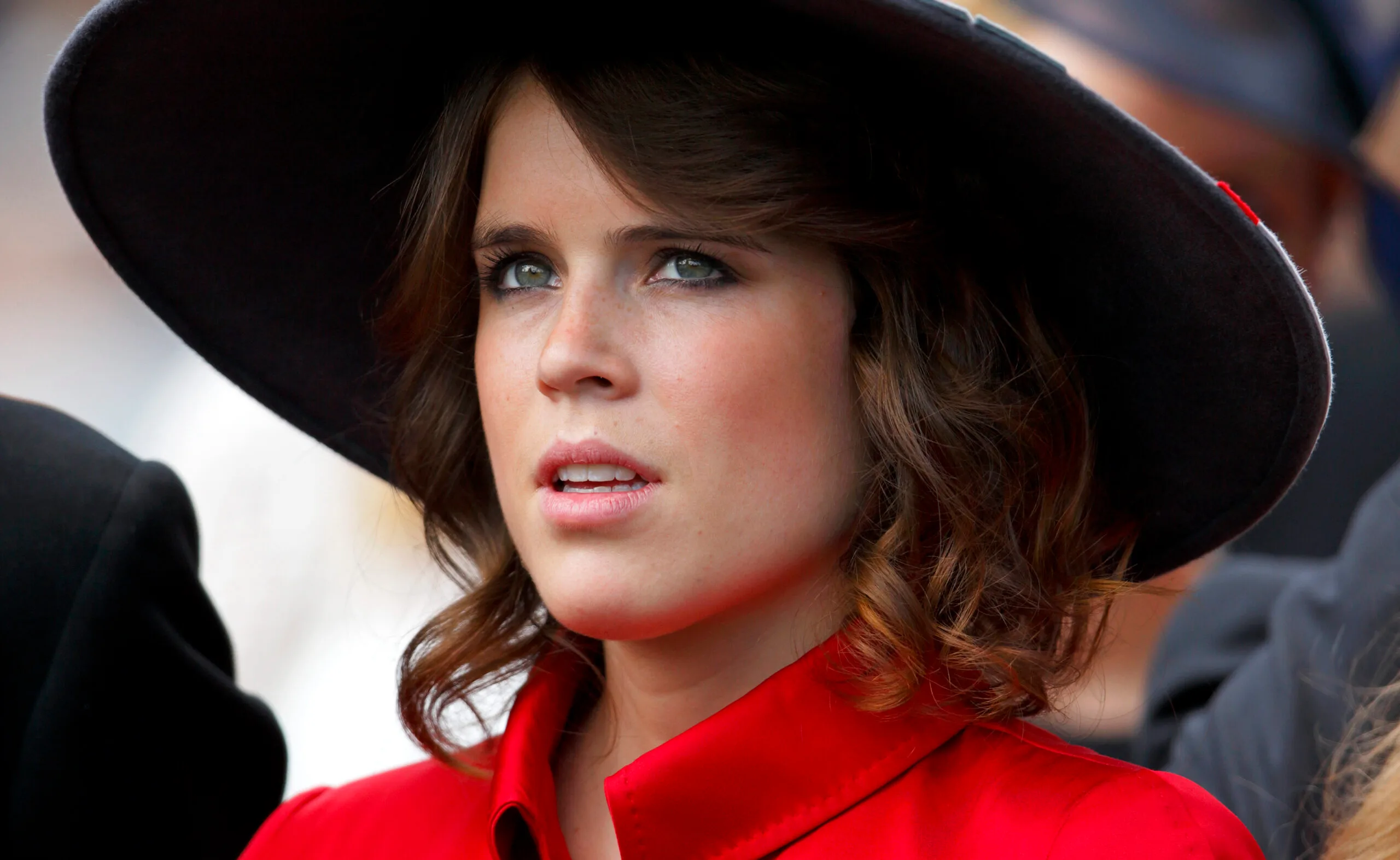 Young woman in a wide-brimmed black hat and red coat, looking forward with a neutral expression.