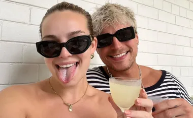 Two people wearing sunglasses smile, one sticking out their tongue, holding a drink against a white brick wall.