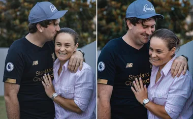 Couple smiling and embracing, displaying an engagement ring outdoors.