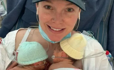 A woman in a cap smiles, holding two newborns with knitted hats in a hospital setting.