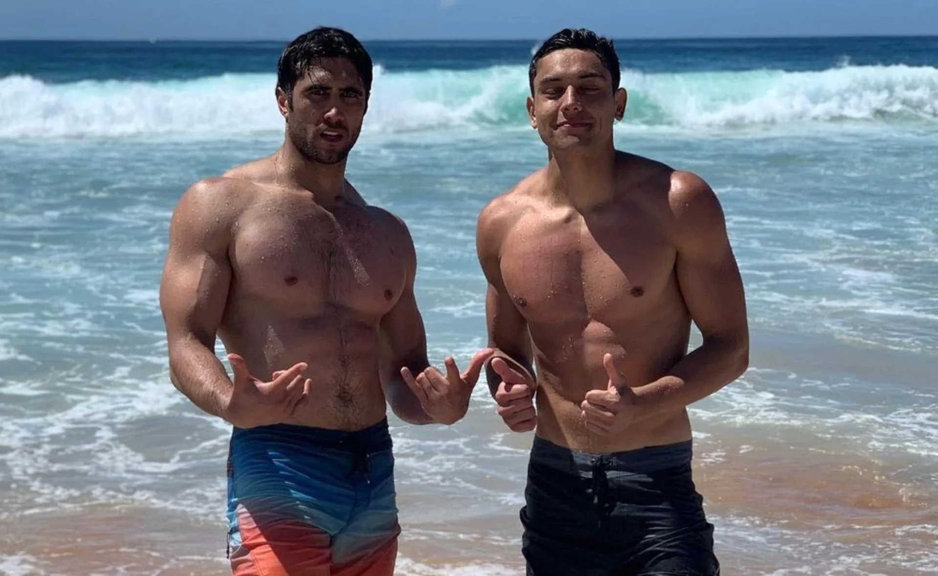 Two men standing shirtless on a beach posing with hand gestures in front of ocean waves.