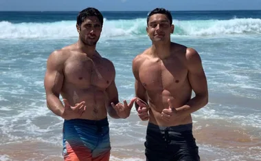 Two men standing shirtless on a beach posing with hand gestures in front of ocean waves.