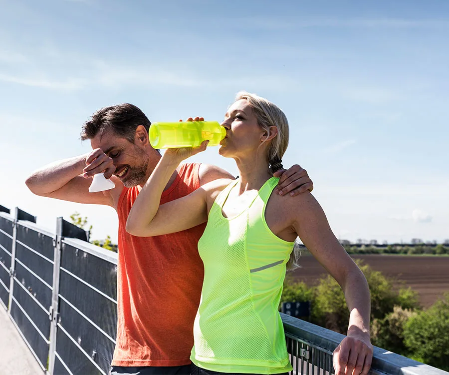 Man and woman taking a break from exercising outdoors, she drinks from a bottle while he wipes sweat.