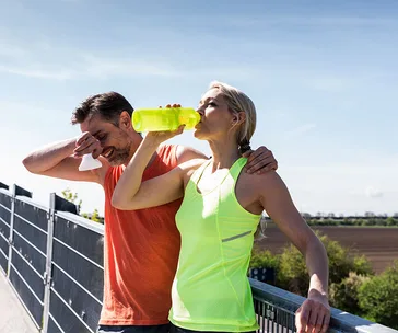 Man and woman taking a break from exercising outdoors, she drinks from a bottle while he wipes sweat.