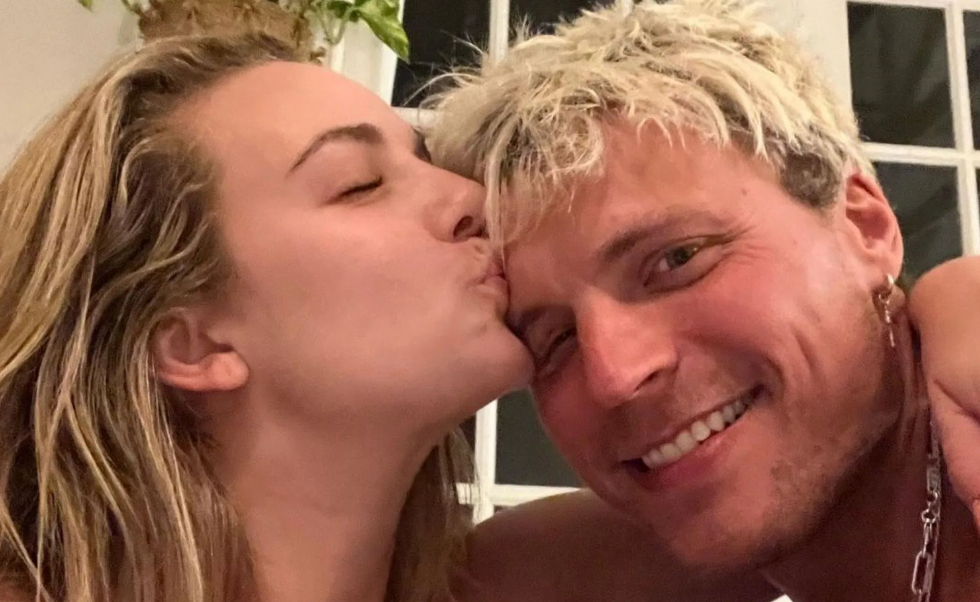 Woman kissing a smiling man on the cheek, indoors with a plant in the background.