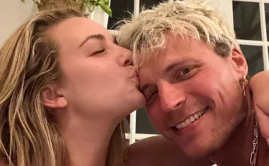 Woman kissing a smiling man on the cheek, indoors with a plant in the background.