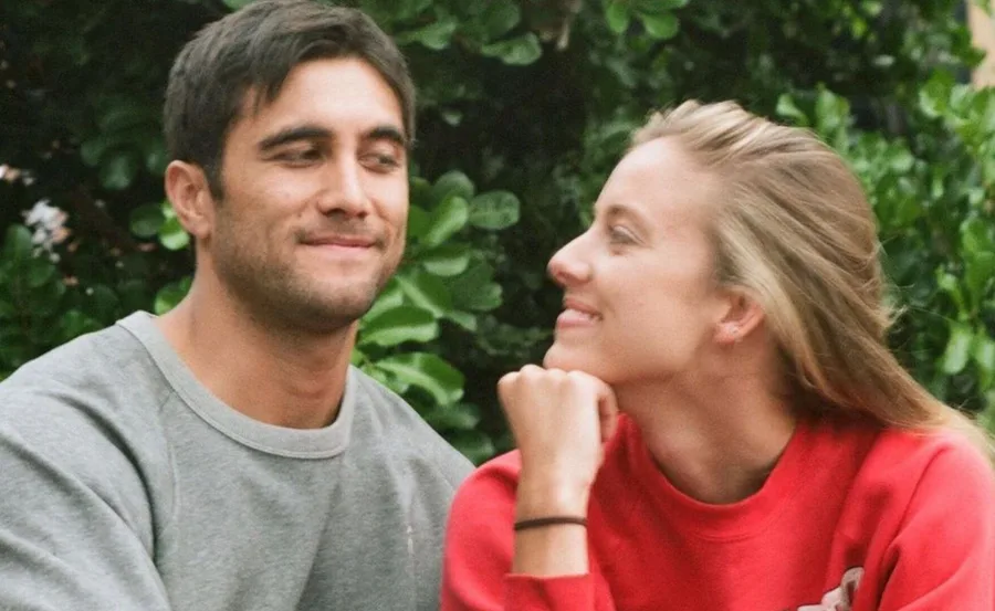 A man in a grey sweater and a woman in a red top exchange smiles, sitting outdoors with green foliage in the background.