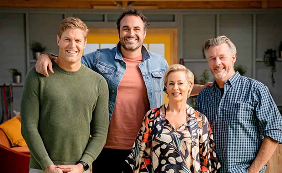 Four people smiling outside a house, casually dressed; the group is in a relaxed setting.