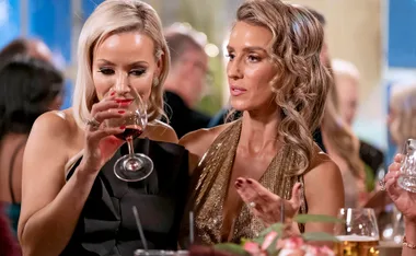Two women at a social event, one sipping wine, the other gesturing, in elegant attire.