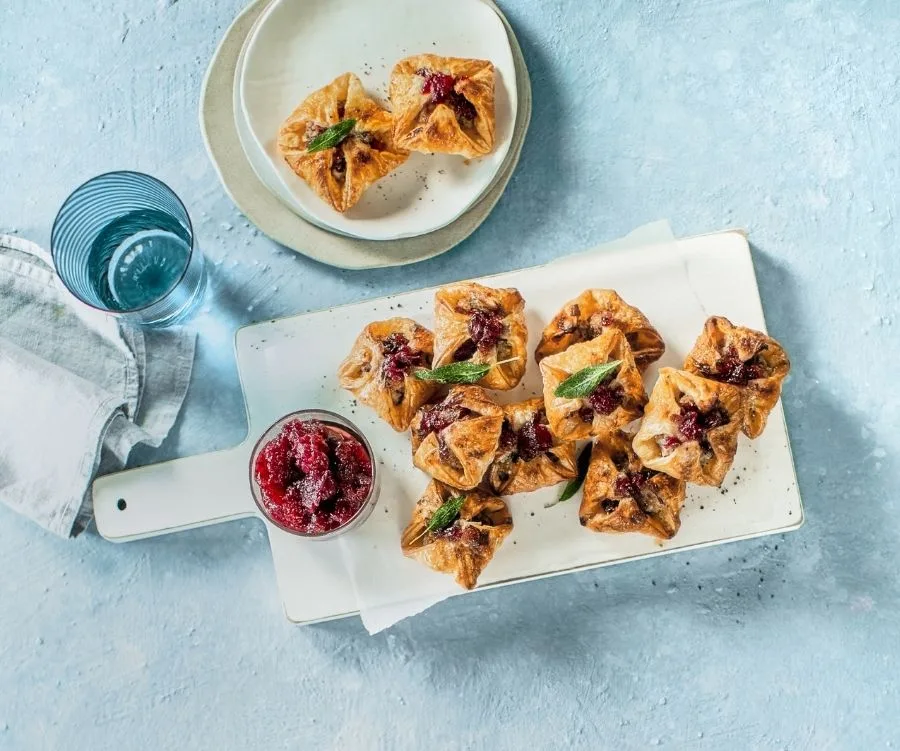 Golden pastry parcels filled with cranberry sauce, arranged on a white board with a glass of water and a napkin nearby.