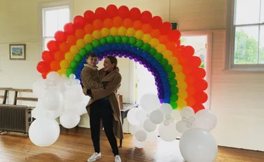 Woman holding a child stands by a vibrant rainbow balloon arch indoors.