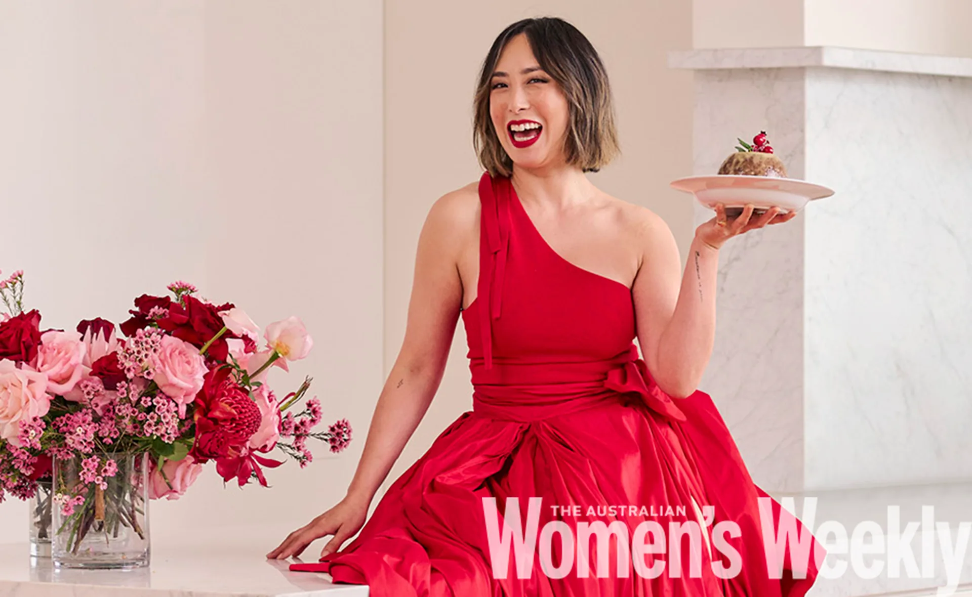 Person in a red dress sits smiling, holding a dessert plate, with pink and red flowers nearby.