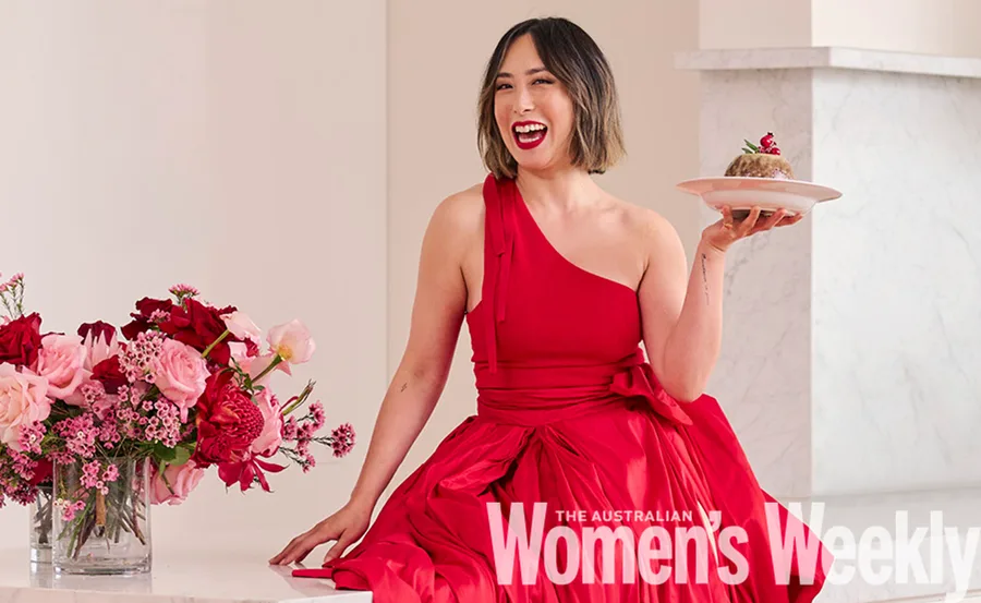 Person in a red dress sits smiling, holding a dessert plate, with pink and red flowers nearby.