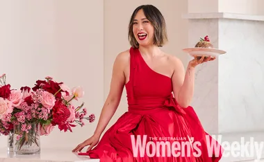 Person in a red dress sits smiling, holding a dessert plate, with pink and red flowers nearby.