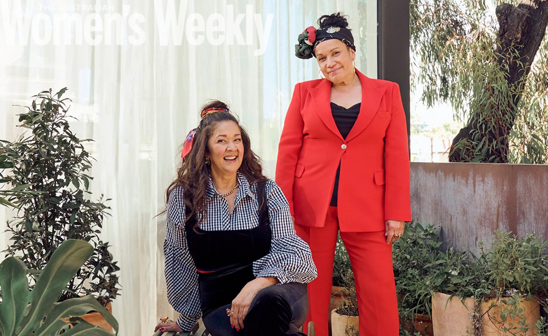 Two women smiling, one seated in black and white attire, the other standing in a red suit, surrounded by plants.