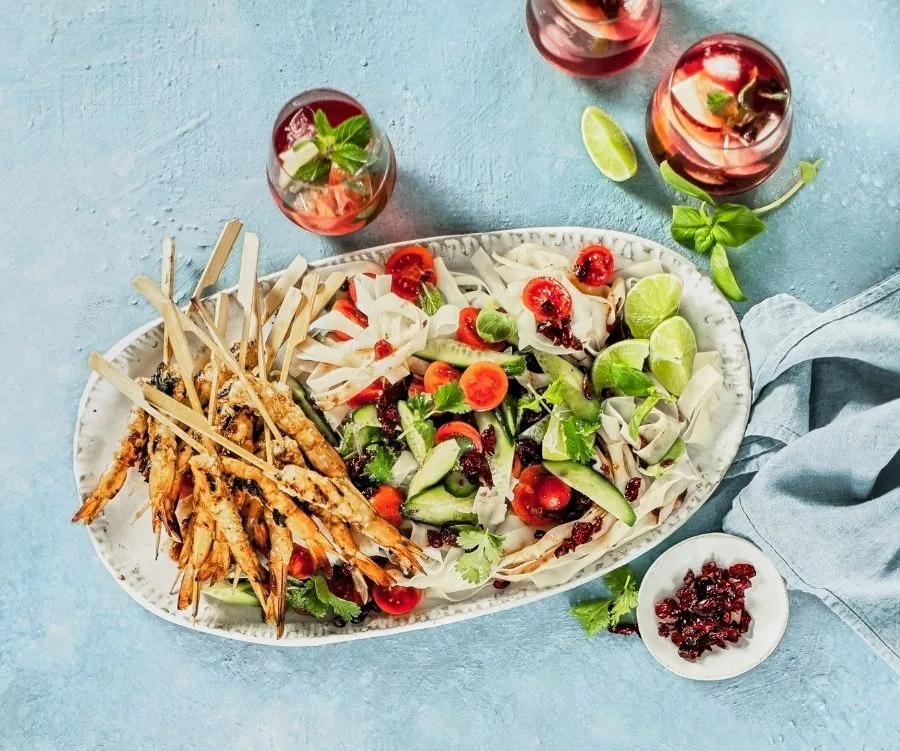 Plate of cranberry-chilli prawns with fresh salad, lime slices, and refreshing drinks on a light blue table.